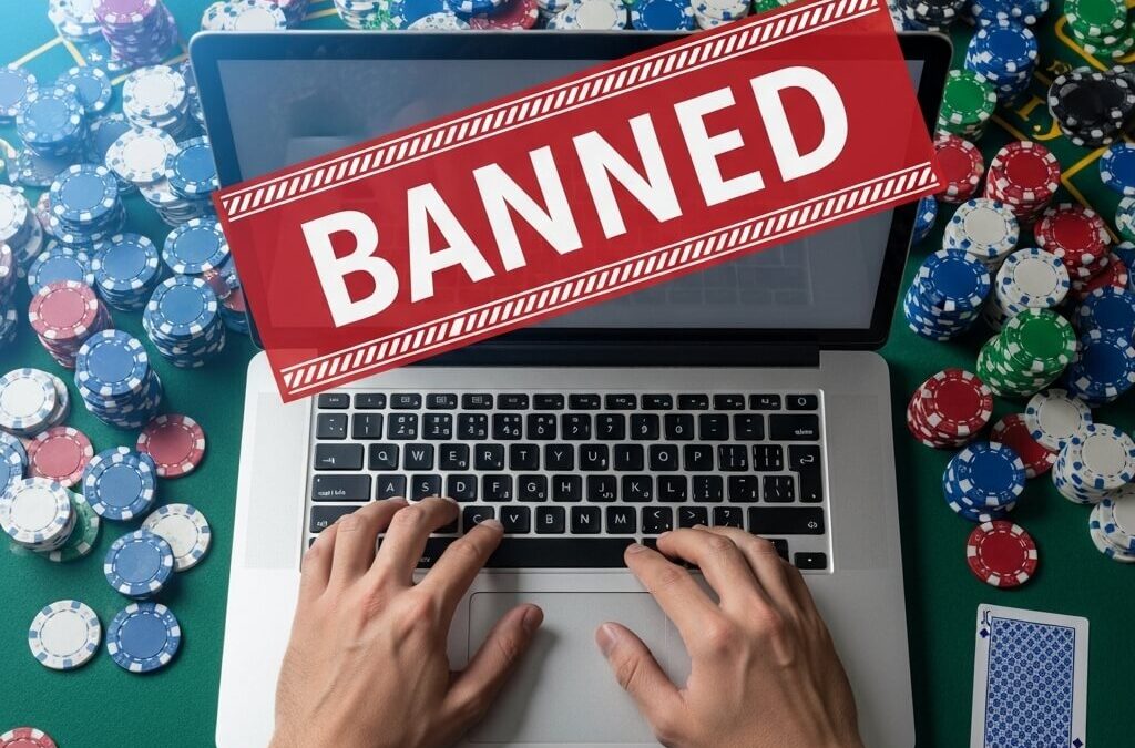 A person at a green felt casino table has been banned from an online gambling site, illustrated by a large red 'BANNED' notice on their laptop screen, which is surrounded by piles of casino chips.